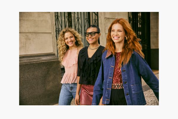 A foto apresenta três modelos. A primeira, mais ao fundo, está com blusa de tule de manga longa com babados rosa bebê e calça jeans. A segunda, do meio, está com blusa de manga longa de tule preto com babados na gola e uma saia de PU cor vinho. A terceira modelo, à frente, veste uma blusa vinho por baixo de uma jaqueta básica.