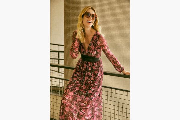 A modelo veste um vestido de manga longa, decote V e comprimento midi. O tecido possui estampa floram com cores sóbrias e finaliza o look com um cinto de PU.