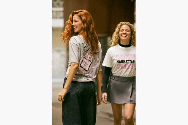 Na imagem tem duas modelos, a primeira está de costas e dá para ver uma estampa grande nas costas da camiseta cinza, finaliza o look com calça jeans. A outra modelo está de frente com uma sobreposição de camiseta branca estampada escrita New York Manhattan sobre blusa de manga longa preta que aparece na gola e mangas abaixo da camiseta. Ela completa o look com shorsaia cinza de alfaiataria.