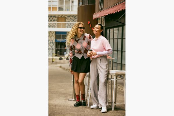 Duas modelos na imagem. A primeira veste um cardigan xadrez estilo retrô com as cores cinza, rosa bebê e vinho. Combinado com saia preta de pregas, meia cano médio vinho e mocassim preto. A outra modelo veste uma camiseta básica oversized sobre uma blusa de frio off de renda que aparece nas mangas. Look completo com calça cinza de alfaiataria com corte amplo e tênis branco.