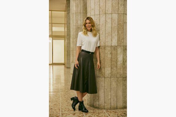 A modelo veste uma saia midi evasê preta confeccionada de PU, tecido que parece com couro. Para finalizar o look, uma blusa off-white básica com manga curta alongada e bota preta de cano curto nos pés.