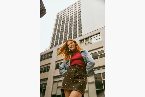 Modelo veste saia de sarja marrom com aplicação de tachas levemente espaçadas em toda a parte da frente da saia. Ela completa o look com uma blusa vermelha básica e jaqueta jeans.