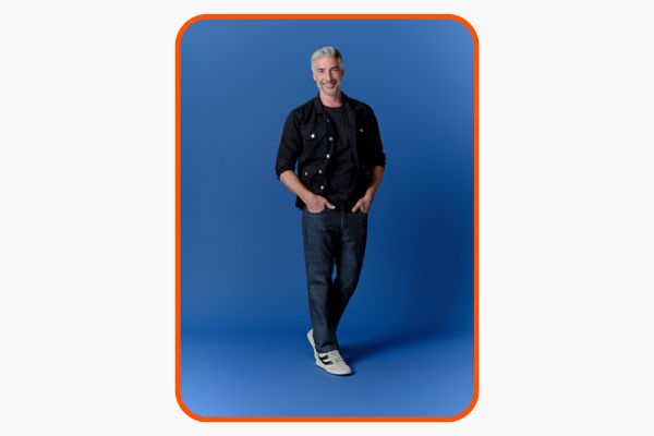 Homem sorridente com camisa preta e jeans sob fundo azul.