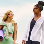 Estilo Athleisure: tendência que traz a versão casual do seu look fitness Duas mulhers com roupa de praticar esporte, mas uma veste uma camisa por cima da roupa fitness e a outra carrega uma esteia de yoga.