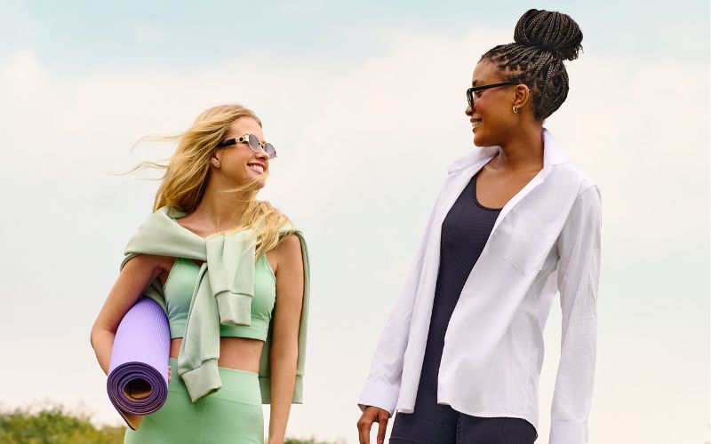 Duas mulhers com roupa de praticar esporte, mas uma veste uma camisa por cima da roupa fitness e a outra carrega uma esteia de yoga.