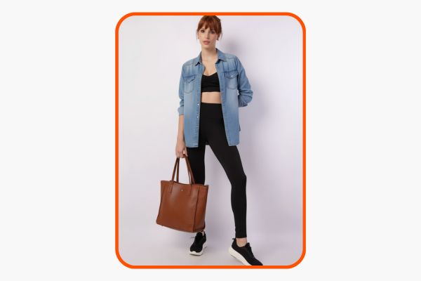 Mulher veste legging e top fitness pretos com uma camisa jeans por cima e usa uma bolsa tote marrom
