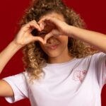 Modelo veste camiseta branca e faz coração com as mãos, o fundo da imagem é vermelho como a cor da campanha de Liquidação da Torra