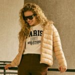 mulher loira veste jaqueta puffer bege sobre camiseta com estampa college.