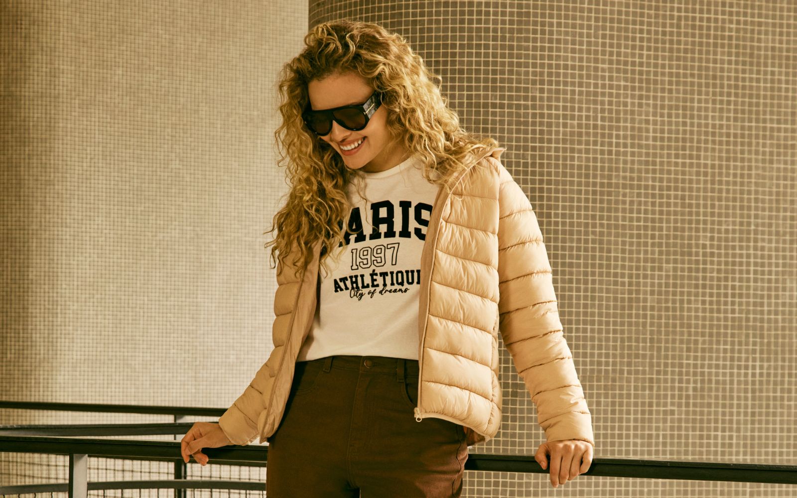 mulher loira veste jaqueta puffer bege sobre camiseta com estampa college.