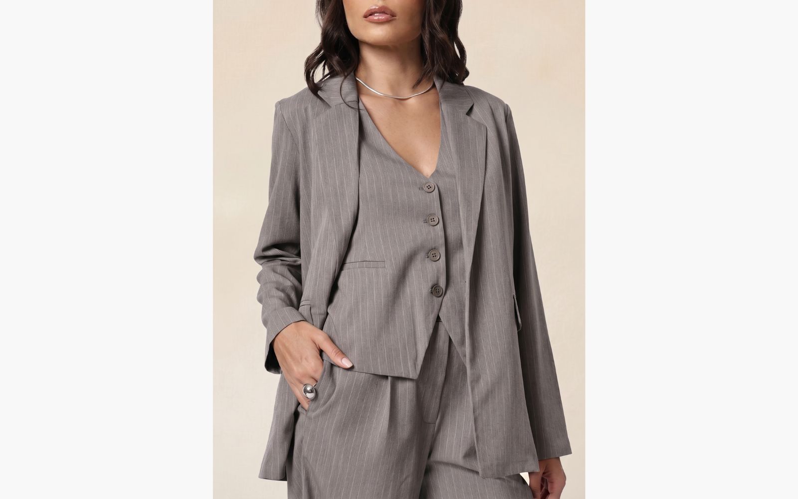 Modelo veste look de alfaiataria composto por: calça social, colete e blazer, todas as peças na cor cinza com estampa risca de giz.