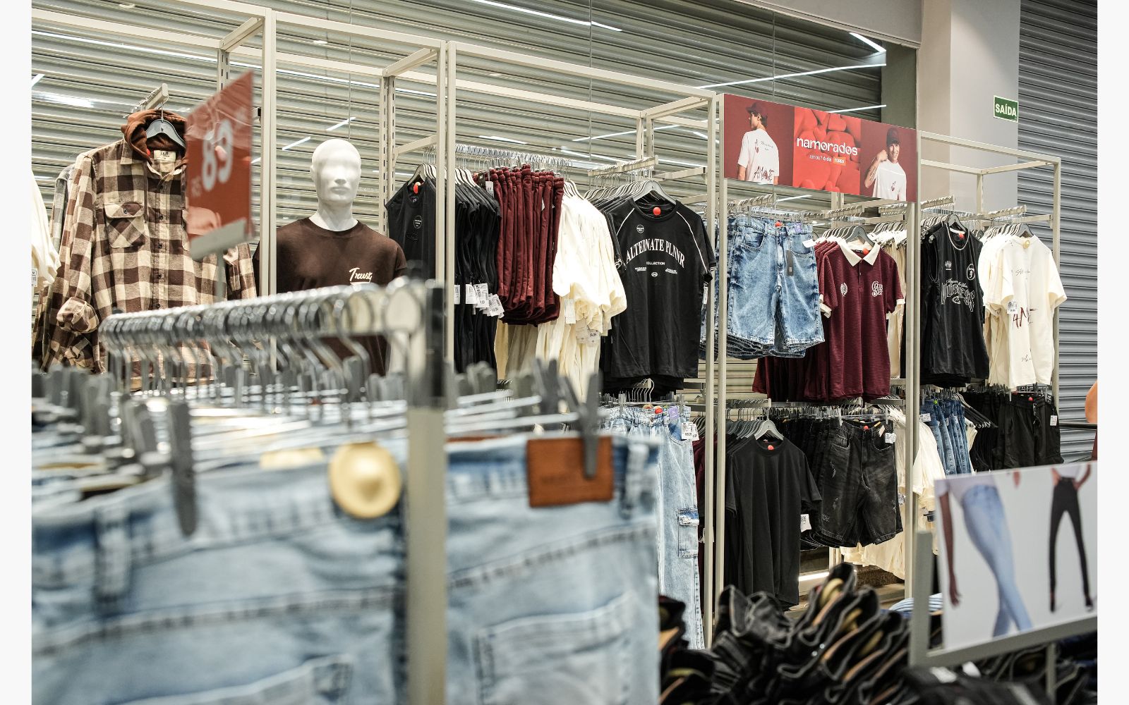 Loja de roupa com roupas em exposição: camisa xadrez de flanela, camiseta oversized com estilo esportivo, bermuda jeans, polo esportiva, calça jeans masculina.