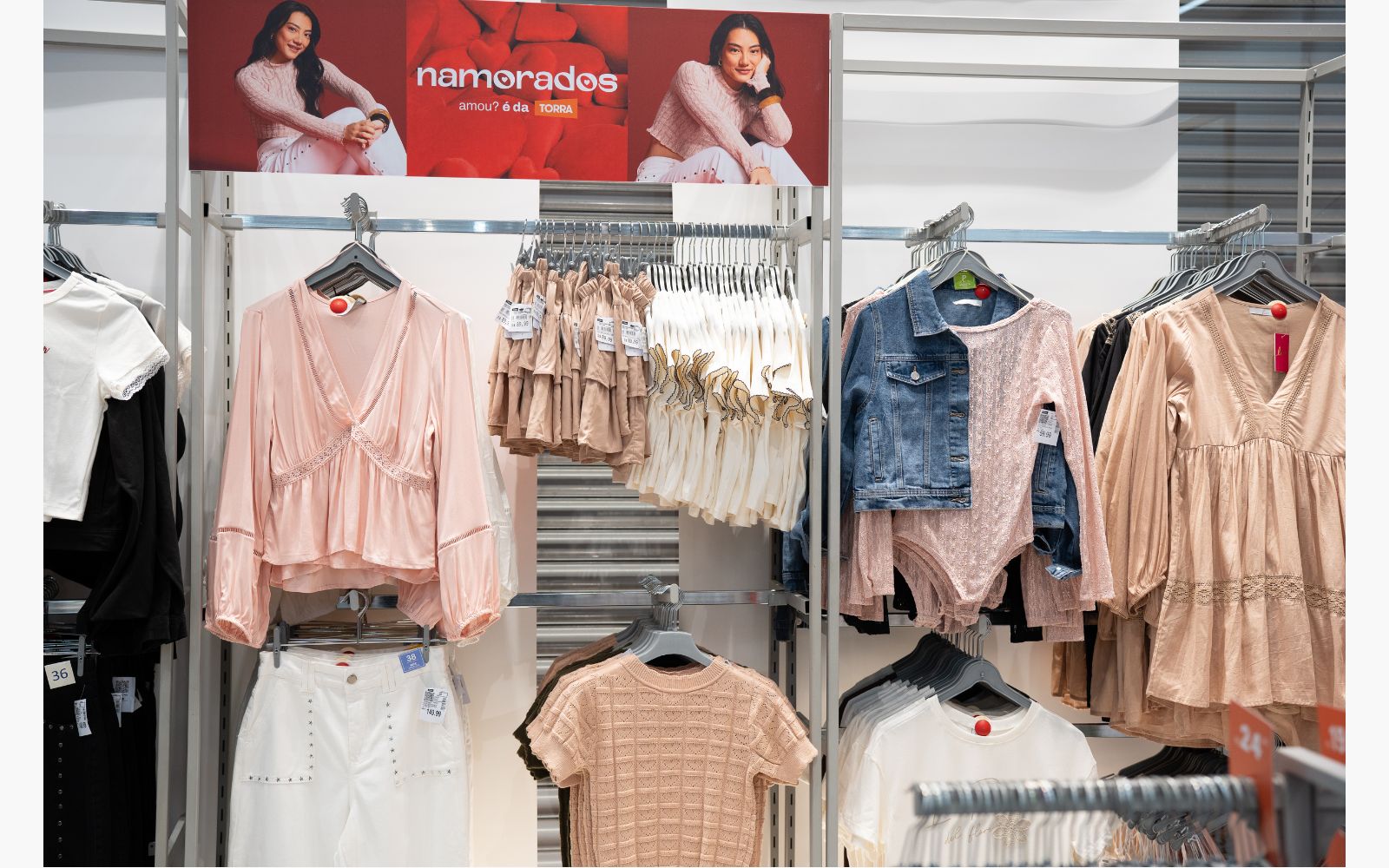 Loja de roupas com roupa feminina em exposição: bata rosé, saia jeans branca com tachas, blusa de tricô nude, body de manda longa e renda, jaqueta jeans cropped, vestido boho chic com rendas.